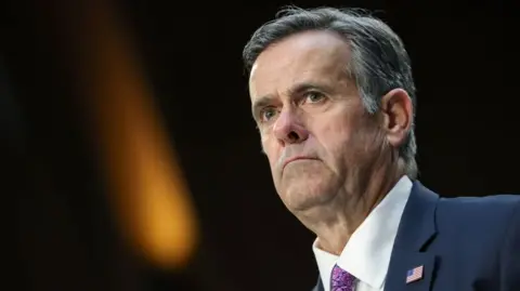 Getty Images A suited John Ratcliffe is wearing a petrol blue suit and mauve tie with a USA flag on his left lapel. The pic is a head and shoulders shot with a microphone visible in front of him as if he is appearing at a hearing.