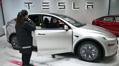 AFP via Getty Images A woman opens the door of a Tesla Model YL electric vehicle as a customer inspects the vehicle