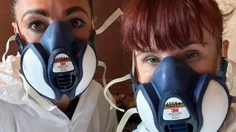 Pocket Rockets Two women in a selfie. The woman on the right has ginger hair tied back and a fringe. Her colleague has brown hair which has been tied back. They are wearing white hazmats suit and navy face masks.