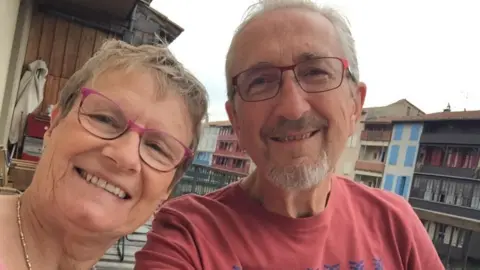 Linda Kitchen Linda and Geoff Kitchen smile together. Both wear rectangular glasses, with Geoff in a red t-shirt. They appear to be sitting on a balcony, with buildings and the cloudy sky visible in the background.
