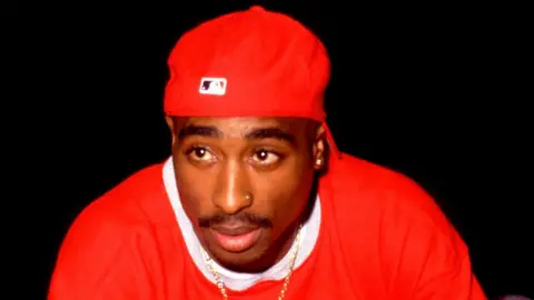 Getty Images Tupac Shakur is seen wearing all red, including a red ball cap, and a gold chain. 