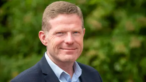 BBC Rhys Hibbert pictured outside in front of a green hedge. He has short fair hair and is wearing a light blue shirt and dark blue jacket.