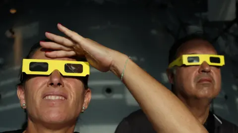 Reuters Two people wearing yellow eclipse-viewing glasses in the foreground look upwards. One person raises a hand above the glasses to shade their eyes. The second person is positioned behind them, also looking towards the sky. The scene appears to be indoors, with a dark, blurred background and sunlight illuminating the faces and glasses.