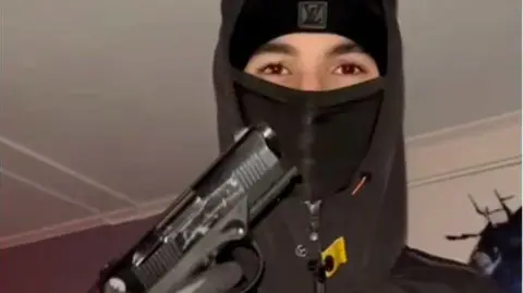 Swedish Police A teenager wearing a black balaclava and hoodie holding up a handgun, with his gloved hand on the trigger