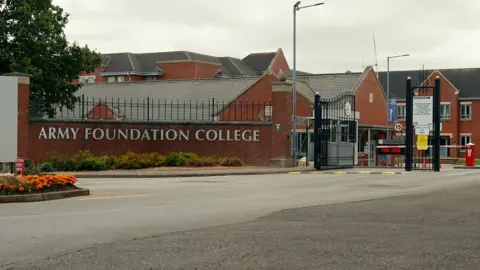 The words Army Foundation College are on a brick wall at the entrance to the campus. Large black gates are open with a red and white barrier slightly beyond.  The college's buildings are behind. 