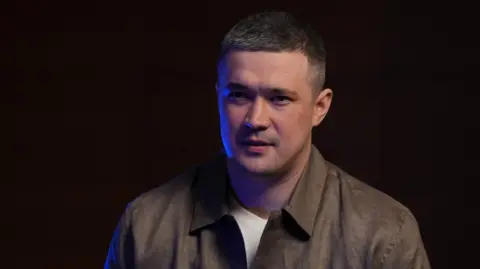 Moose Campbell/BBC A man dressed in a white T-shirt and brown collared shirt in front of a black background sits talking to someone with their back to the camera.