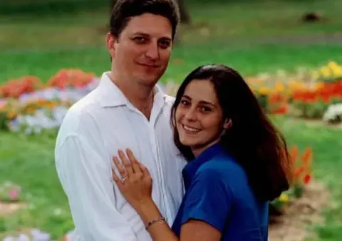 Tom Resta John Resta is on the left, he has dark hair and is wearing a white linen shirt and his wife Sylvia is on the right, with dark long hair and a blue short-sleeved shirt. She is pressed against him, her left hand on his chest. It has a wedding ring on it. They are both smiling. Behind them is grass with flowers. 