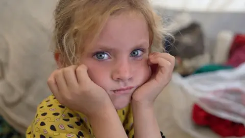 A girl with in a yellow outfit stares into the camera.