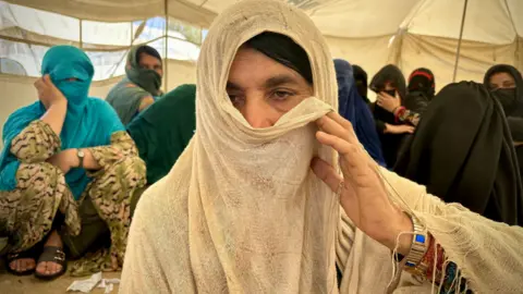 Zarina, an Afghan returnee, inside a tent with several others is using her veil to partially cover her face as she faces the camera. 