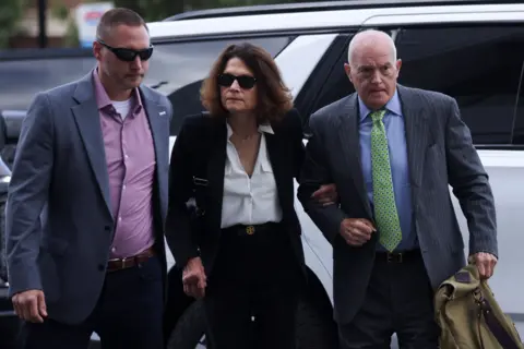 Reuters Charlie Kirk's mother, Kathryn, wears sunglasses and a dark suit over a white blouse as she holds the arm of her husband, Robert, wearing a pinstripe suit and green tie while they arrive at the Utah courthouse on Tuesday