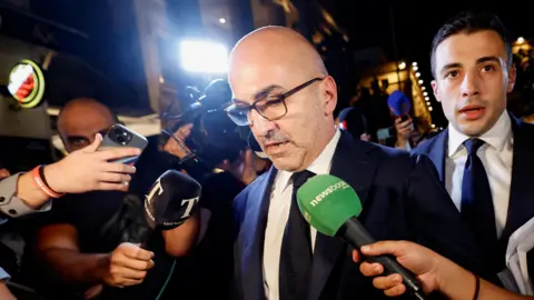 Reuters Yorgen Fenech wears a black suit and glasses surrounded by journalists