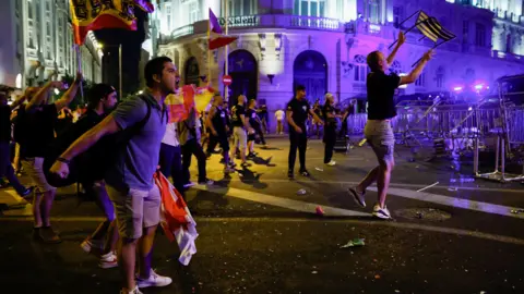 Reuters A man shouts at police with a barricade on the right and police lights visible. Other protesters wave flags. 