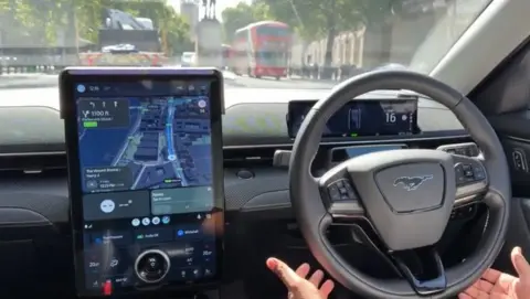 A passenger's view inside a robotaxi as it drives through London, with the hands of the safety driver not touching but around the wheel of the car, and a large black tablet with a map on it showing where the car is going.
