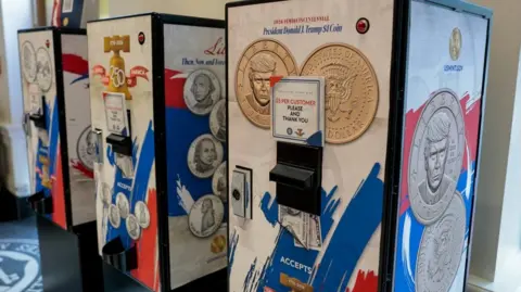 Reuters Three vending machines selling $1 coins featuring President Donald Trump's image at the US Mint Coin Store in Washington DC. On each machine there is a sign saying "$5 per customer please and thank you".