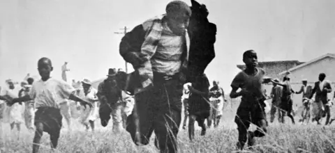 Universal History Archive/UIG via Getty images A grainy black & white image of a group of people running towards the camera. In the front is a man holding up his jacket - just behind him, either side, are tow children.
