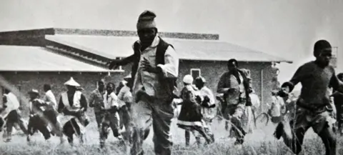 Universal History Archive/UIG via Getty images A grainy black & white image of a group of people running towards the camera. A low-rise building can be seen behind them. 