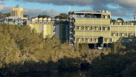 Supplied A large green and grey building situated amid bushland and a river down below.
