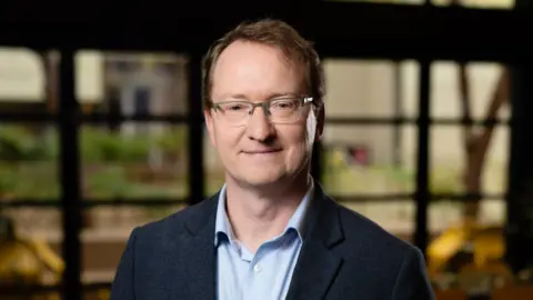 Adrien Rossignol A middle aged man with short brown hair and glasses wearing a blue shirt and jacket smiles at the camera