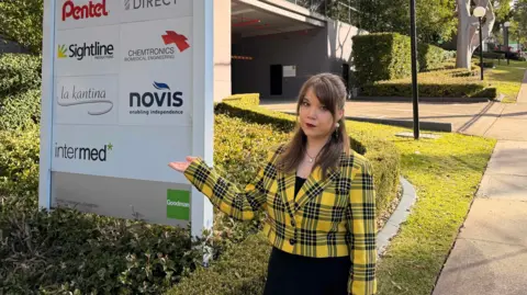 Supplied by Rochelle Flood A woman dressed in a yellow and black checked blazer gestures with her hand to a sign in front of a building complex. A green lawn and a gum tree are in the background.