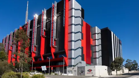 Getty A huge red and grey windowless building with pipes on the outside against a blue sky.