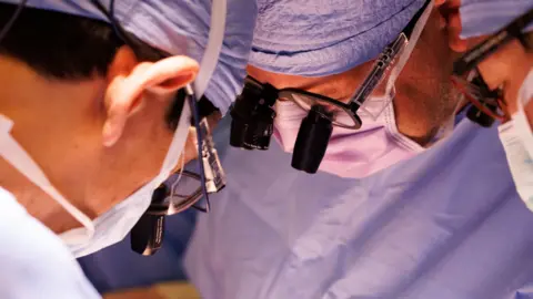 Kate Flock/Massachusetts General Hospital A tightly framed photograph shows several members of a surgical team gathered closely during a procedure. Each person is wearing blue surgical clothing, face masks, and magnifying lenses mounted on protective eyewear. The image focuses on their faces and equipment as they lean inward and look down toward an area outside the frame. Bright operating-room lighting illuminates the scene, while the background remains softly blurred in shades of blue. The composition emphasises concentration, precision, and the collaborative nature of a medical procedure.