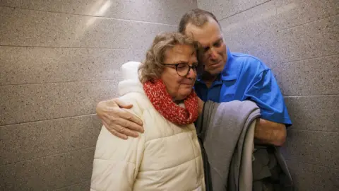Kate Flock/Massachusetts General Hospital The image shows two adults standing together in a narrow area with grey stone or ceramic tiled walls. One person is wearing a bright blue polo shirt and holding a folded grey garment, while the other is dressed in a light-coloured padded winter coat with a thick red knitted scarf and glasses. The two individuals are positioned very close to one another, with an arm wrapped around the other's shoulder. The composition is tightly framed around the pair, drawing attention to their physical closeness.
