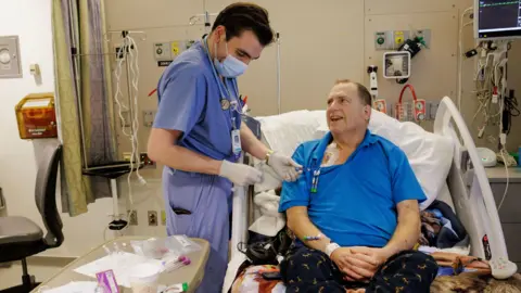 Kate Flock/Massachusetts General Hospital The image shows a hospital room with a patient seated in an adjustable bed, wearing a bright blue polo shirt and patterned dark trousers. A healthcare worker dressed in blue scrubs, gloves, a face mask, and a stethoscope is standing beside the bed and handling tubing connected to a medical access point on the patient's upper chest. Various items including syringes, medical packaging, paperwork, and a disposable cup are arranged on a bedside table in the foreground. The room contains hospital equipment such as wall-mounted oxygen and suction outlets, monitoring devices, intravenous poles, and cables connected to equipment near the bed.