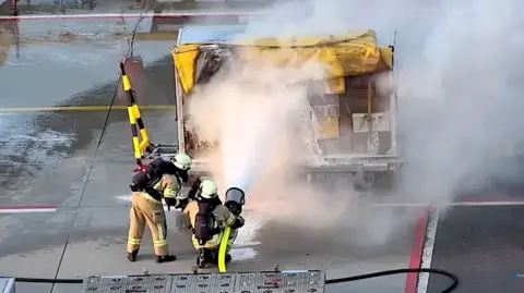 Police handout Two fire officers are spraying foam on an airport cargo trolley. Smoke is spreading from the trolly. 