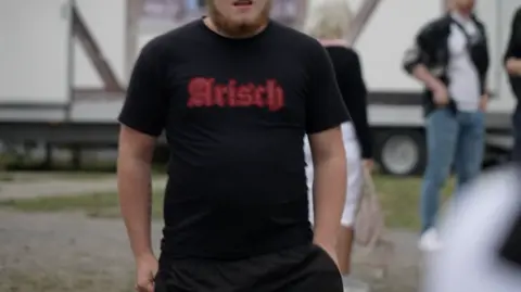 The torso of a man wearing a black t-shirt which says 'Arisch', German for Aryan written in a red Gothic-style font.