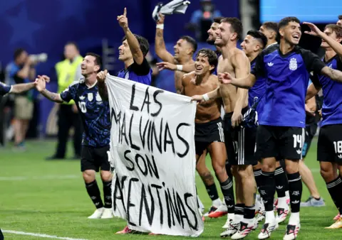 REUTERS/Amanda Perobelli Football players, some with their shirts off, stand or jump while smiling on a green football pitch. Two of them hold a white banner that says in black writing "Las Malvinas Son Argentinas" - the Falklands are Argentina's.