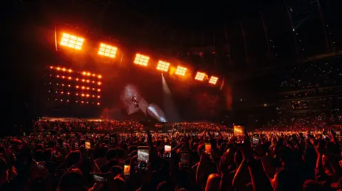 Roc Nation Jay-Z performs on stage as a sea of fans watch along. There are phones raised in the air and there is a red hue cast over the crowd.