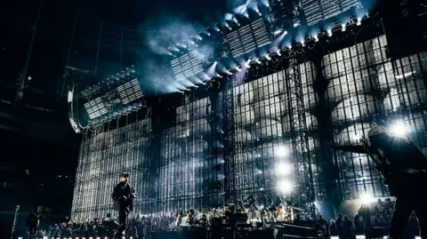 Roc Nation Jay-Z performs on stage in front of a large LED screen. There is an orchestra band behind him. Jay-Z wears all black.