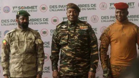 Reuters Mali's Assimi Goita, Niger's Gen Abdourahamane Tiani and Burkina Faso's Captain Ibrahim Traore - all in military uniform - pose together for photographs. They're standing in front of a board that says, in French, that this is the first heads of state summit of the Alliance of Sahelian States. 