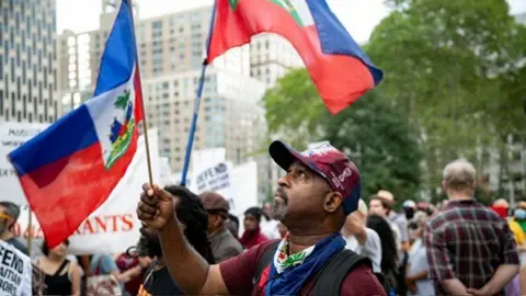 Reuters New York City skyscrapers and trees can be seen in the background as a man stands in a crowd of people waving placards; he wears a baseball cap, scarf and waves a Haitian flag