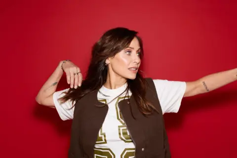 Rankin Natalie Imbruglia poses in a brown puffer jacket and a t-shirt with the logo "1989". She is standing in front of a red backdrop with her arms outstretched
