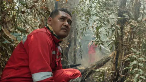 Ahmad Dhani for BBC A man in red firefighting uniform crouches in bushland