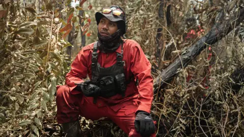 Ahmad Dhani for BBC A man wearing red firefighting uniform, tactical vest, gloves, neck gaiter and hat crouches in a forest, holding a handheld radio device