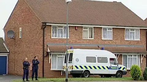 A still from a video showing a small, mid-century semi-detached home in Birmingham with net curtains and a satellite dish. A police van is parked outside, blocking the front door, and two police officers stand to the side of the building, with their arms folded