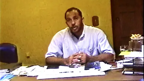 SDNY A man wearing a lilac shirt sits at a desk covered with paperwork. 