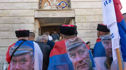 Reuters Men wearing flags as capes, with a picture of former Bosnian Serb wartime general Ratko Mladic, stand amongst people gathered at the Church of St. Luke, where the body of Mladic lies in state before burial, in Belgrade, Serbia, September 7, 2026. 