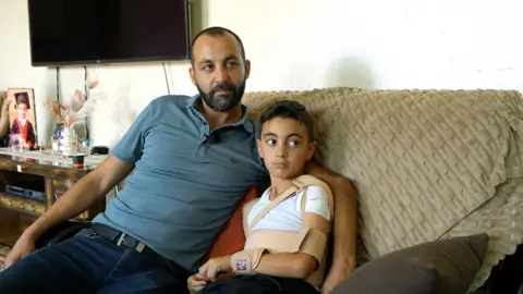 Salim sits on a sofa with his father. His arm is in a sling. 
