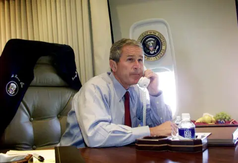 Getty Images President Bush is at a desk on the plane with his jacket off and hung on the back of his seat, he is wearing a light blue shirt and maroon tie. He has a phone in his left hand and seems to be in conversation.