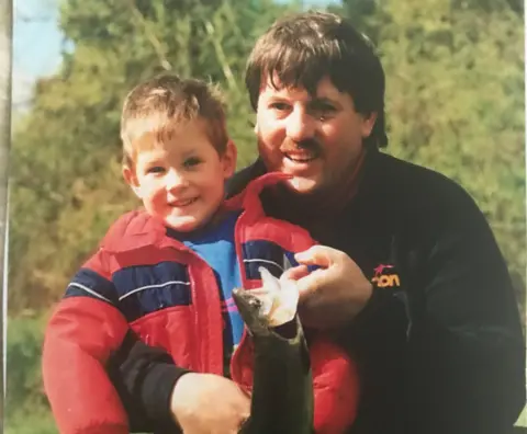 Brett Eagleson Brett is on the left and a young boy in this undated photo. He is wearing a red jacket and he's smiling at the camera. He is in the arms of his dad Bruce who is wearing a black jacket and is holding a fish in his left hand.