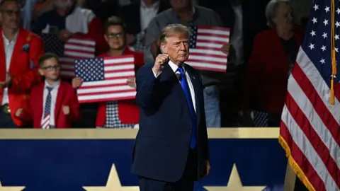 Getty Images President Trump, in a blue suit and tie" raises a fist on stage. Behind him, people are gathered and holding American flag signs. 