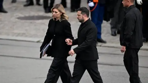 Reuters Olena and Volodymyr Zelensky hold hands as they leave the funeral. They are both dressed in black and are outside.