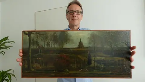 Arthur Brand A man with glasses and wearing a blue shirt stands holding a painting up to the camera. The camera is quite dark and shows a figure walking through a garden, with a church-like building in the background.