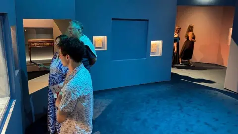 Jeremy Audouard/AFP via Getty Images People stand looking a glass display case on the left of a room, which has a blue carpet and blue walls. There is a wall with a blank square where there is nothing in it.