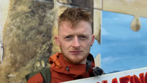Man with blonde hair, red coat holding a sign.