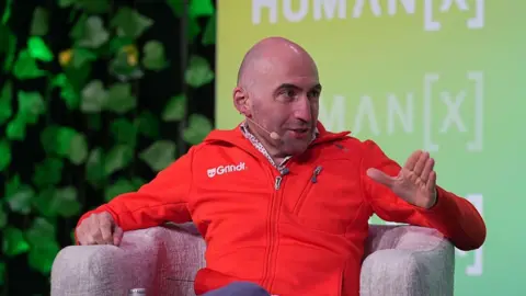 Getty Images George Arison speaks onstage during the "AI, Identity, and the Future of Connection" panel at the HumanX Conference San Francisco 2026 . He wears a red casual zip up sweater with the Grindr logo in white and speaks into a microphone. 