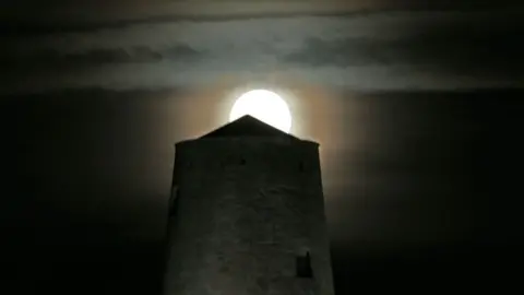 Getty Images A bright full moon rises above a castle tower made of grey stone with a small peaked roof. There are some layers of cloud in the distance of a dark black sky. 